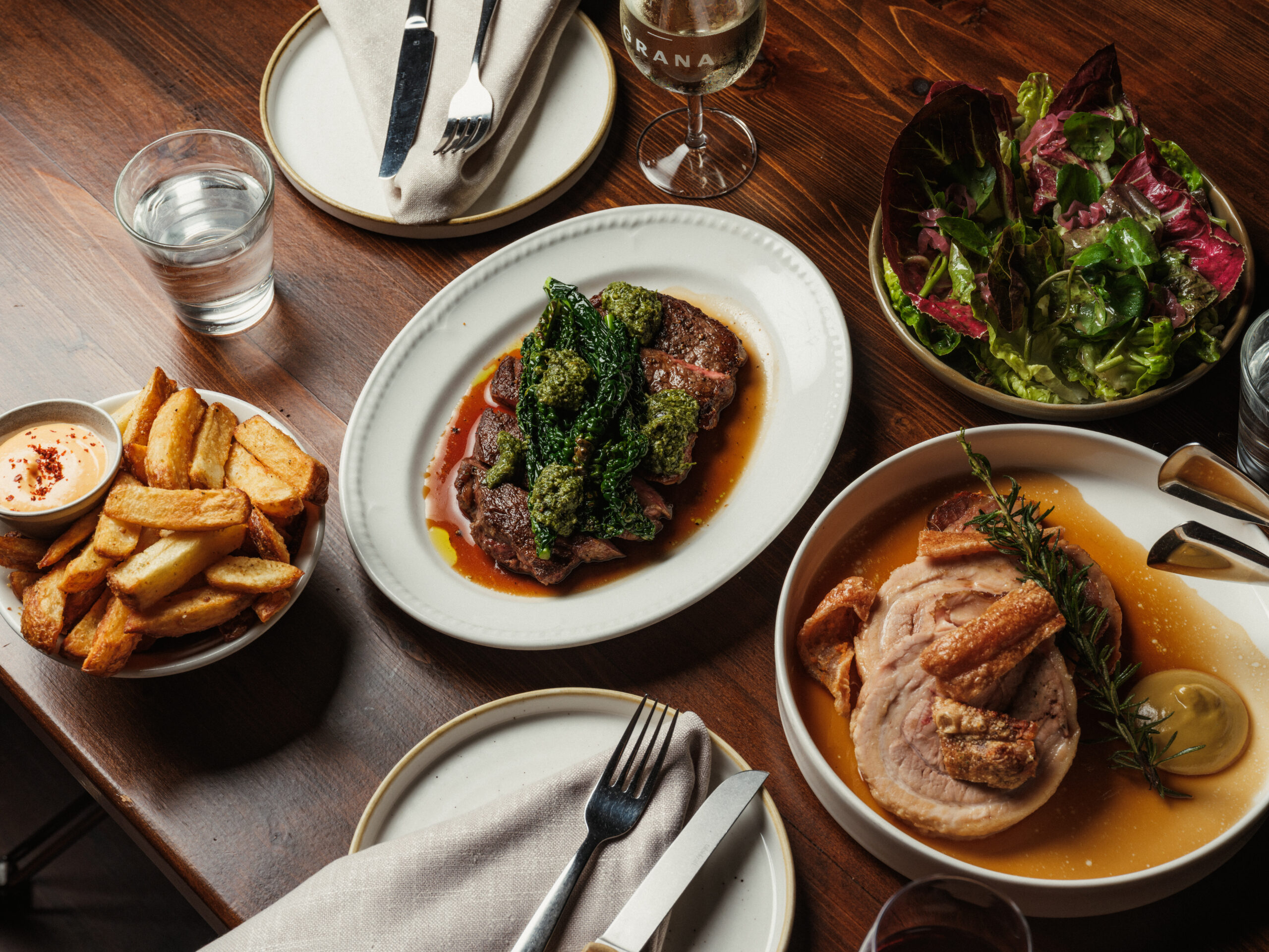 Shared meat, salad, fries, and wine on a timber table at Grana